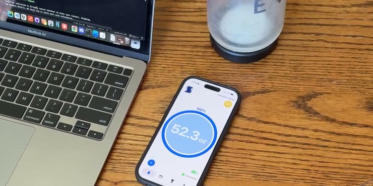 A water bottle with a Sponge tracker at its base beside a laptop, next to a phone showing 52.3 oz logged in the Sponge app